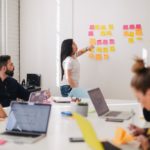 People sitting in a meeting room while a woman at a board in the front points toward some sticky notes on the board.