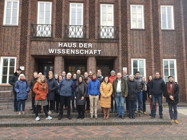 A group of people looking towards the camera standing in front of a brick building on which it says "Haus der Wissenschaft".