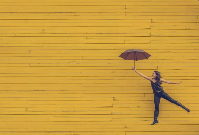 A young woman with brown hair holding an umbrella. She seems like flying with it. The background looks like yellow wooden planks.