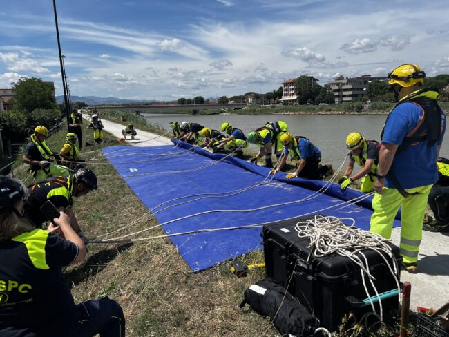 Menschen in Sicherheitskleidung rollen gemeinsam ein blaues Banner auf, welches auf einem Damm an einem Fluss liegt. Weitere Personen in Sicherheitskleidung halten Seile, welche über das Banner gelegt sind.